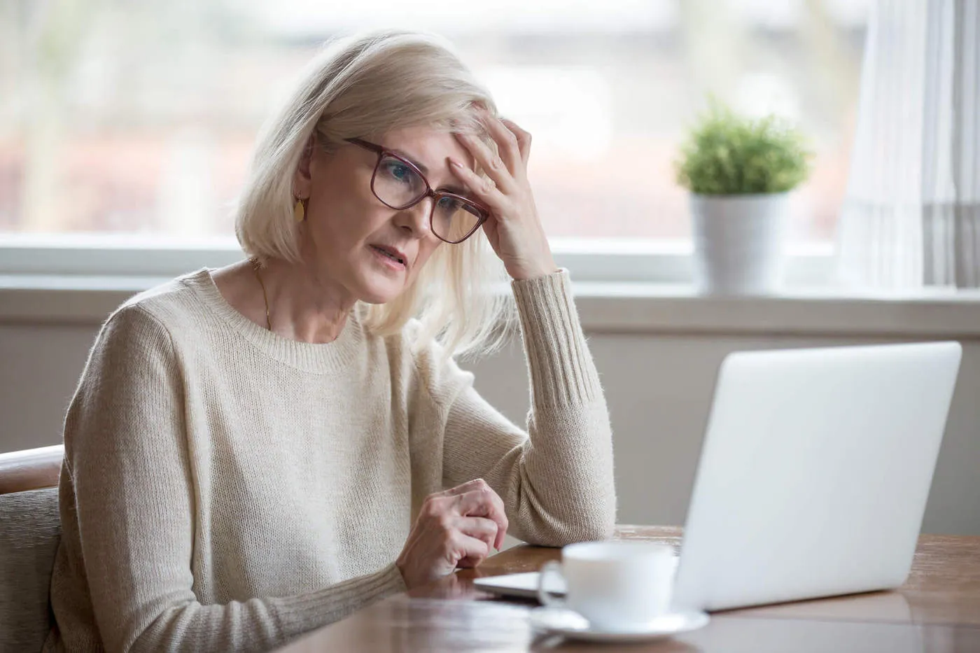 An elderly woman put her hand on her forehead as she looks at her computer.