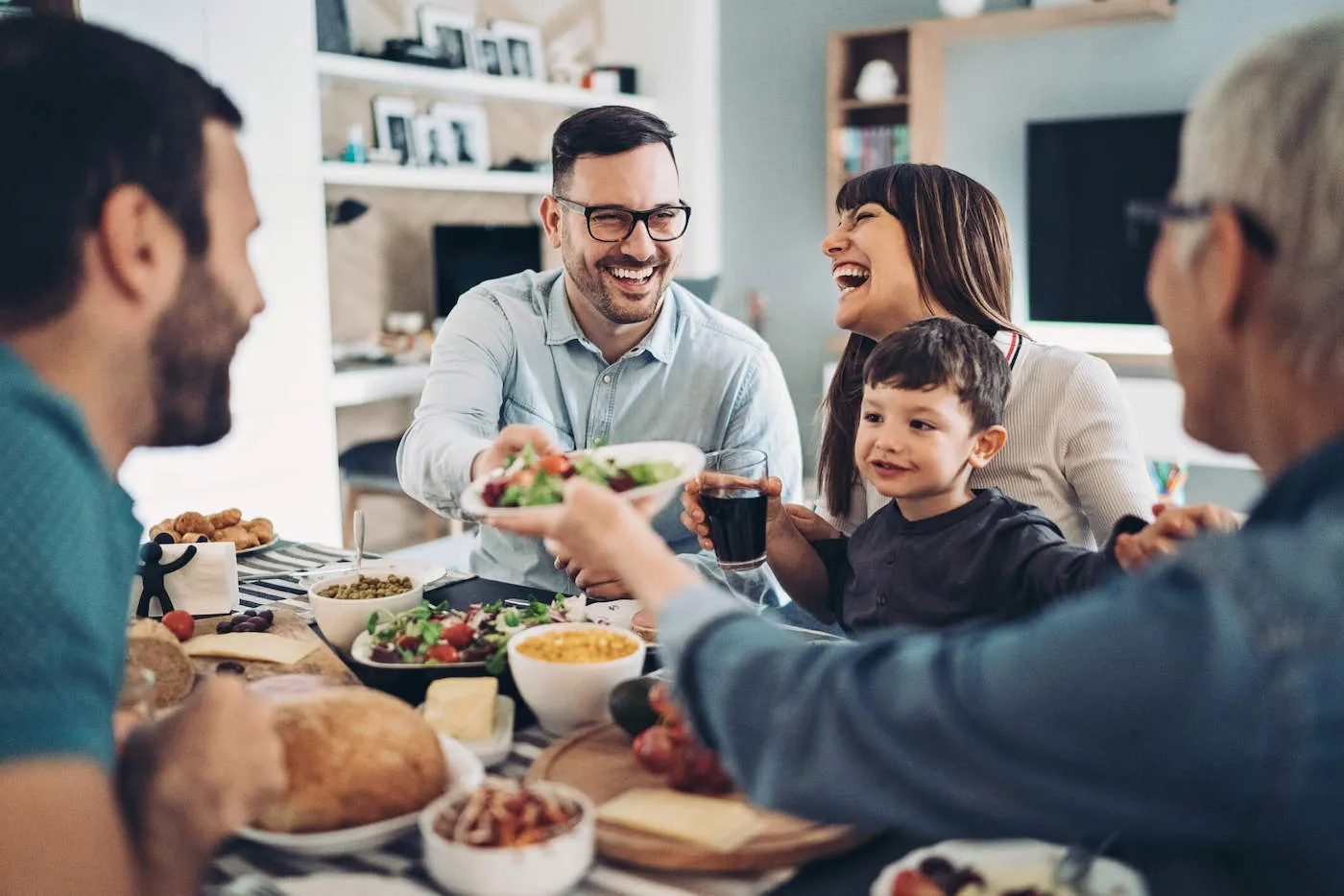 A family of five are sharing a meal at they kitchen table while they are laughing and talking with one another.