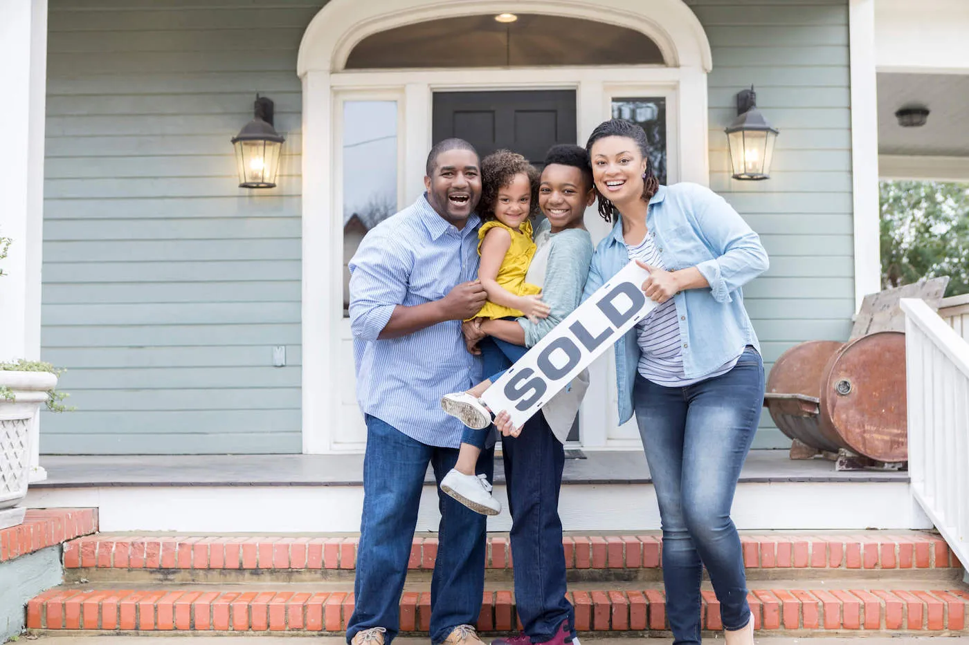 A family of four smile while holding a "sold" sign in front of their new home.