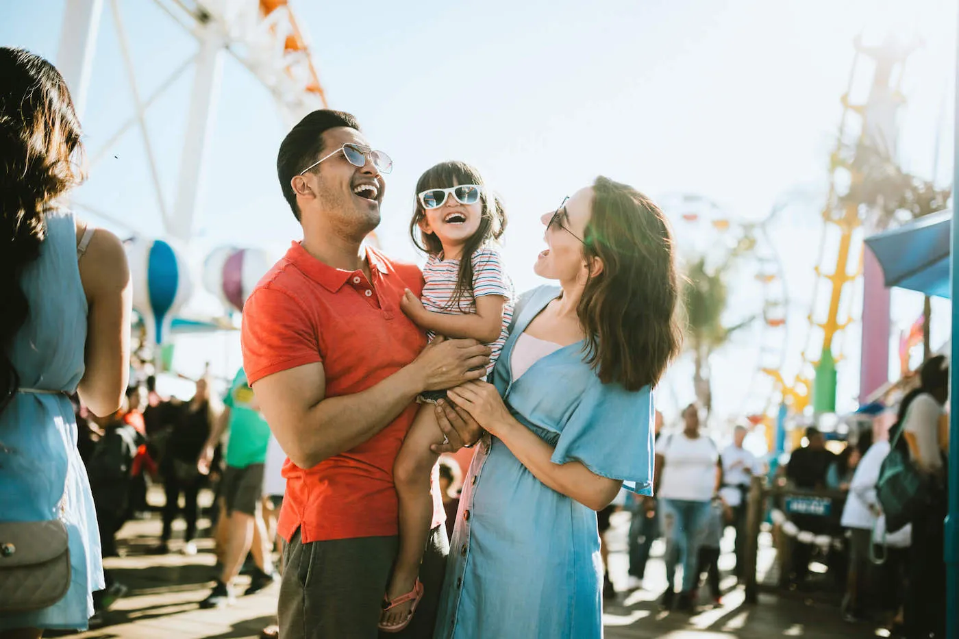 Mom and dad are happily holding their daughter as they all wear sunglasses at the amusement park.