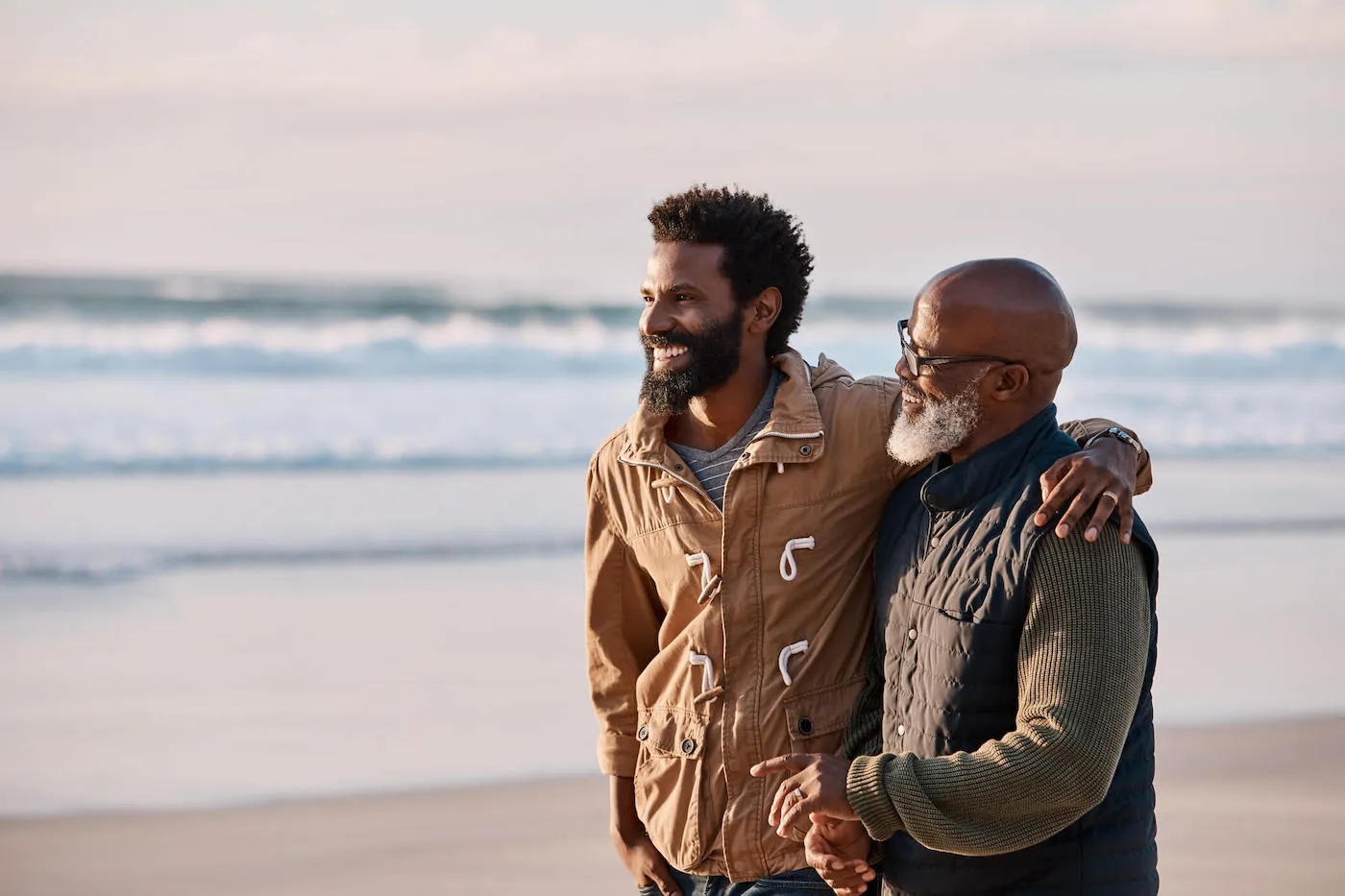 A father and son walk on the beach together, smiling, while the son has his arm around his father.