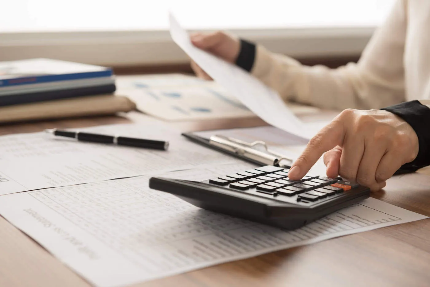 A person wearing a white sweater uses a calculator while documents are laid out on the table.