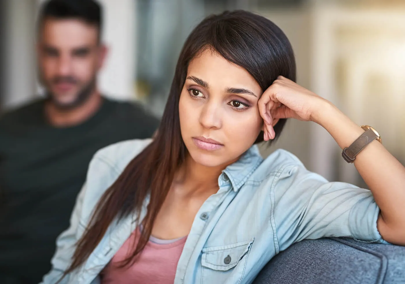 A woman sitting on a couch staring wistfully into the distance. A man is sitting behind her.