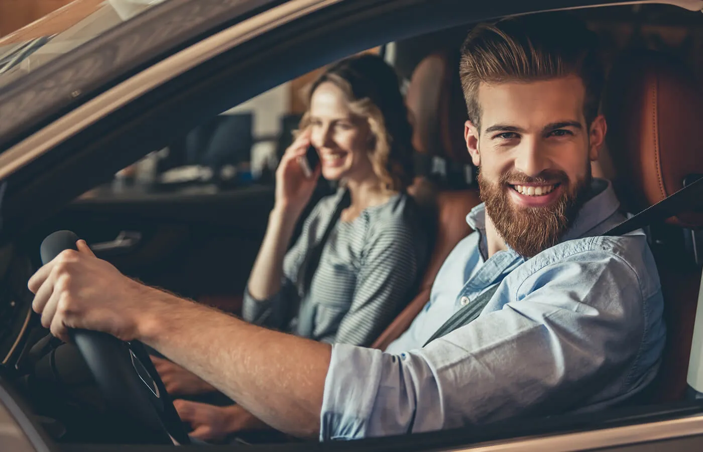 A man driving a car with a woman passenger in the front seat. He is smiling and looking out the driver's side window.