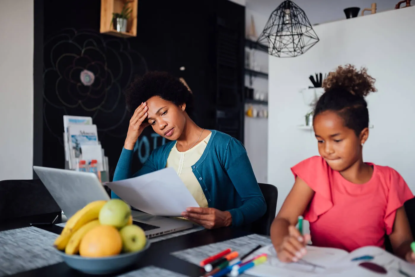 A mother looks at a document next to her daughter that is drawing.