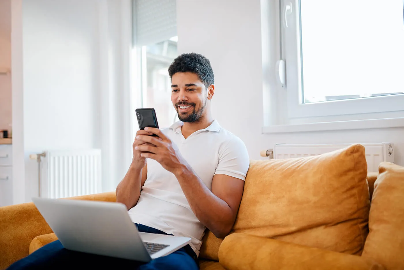 A young man sits on an orange couch while smiling and using his cellphone.