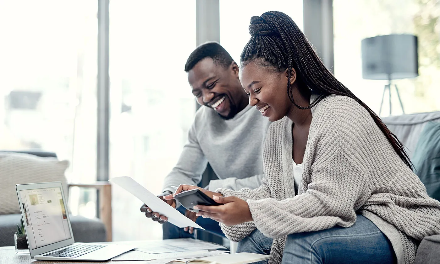 A couple sit on the couch together and smile as they look over documents on the living room table.