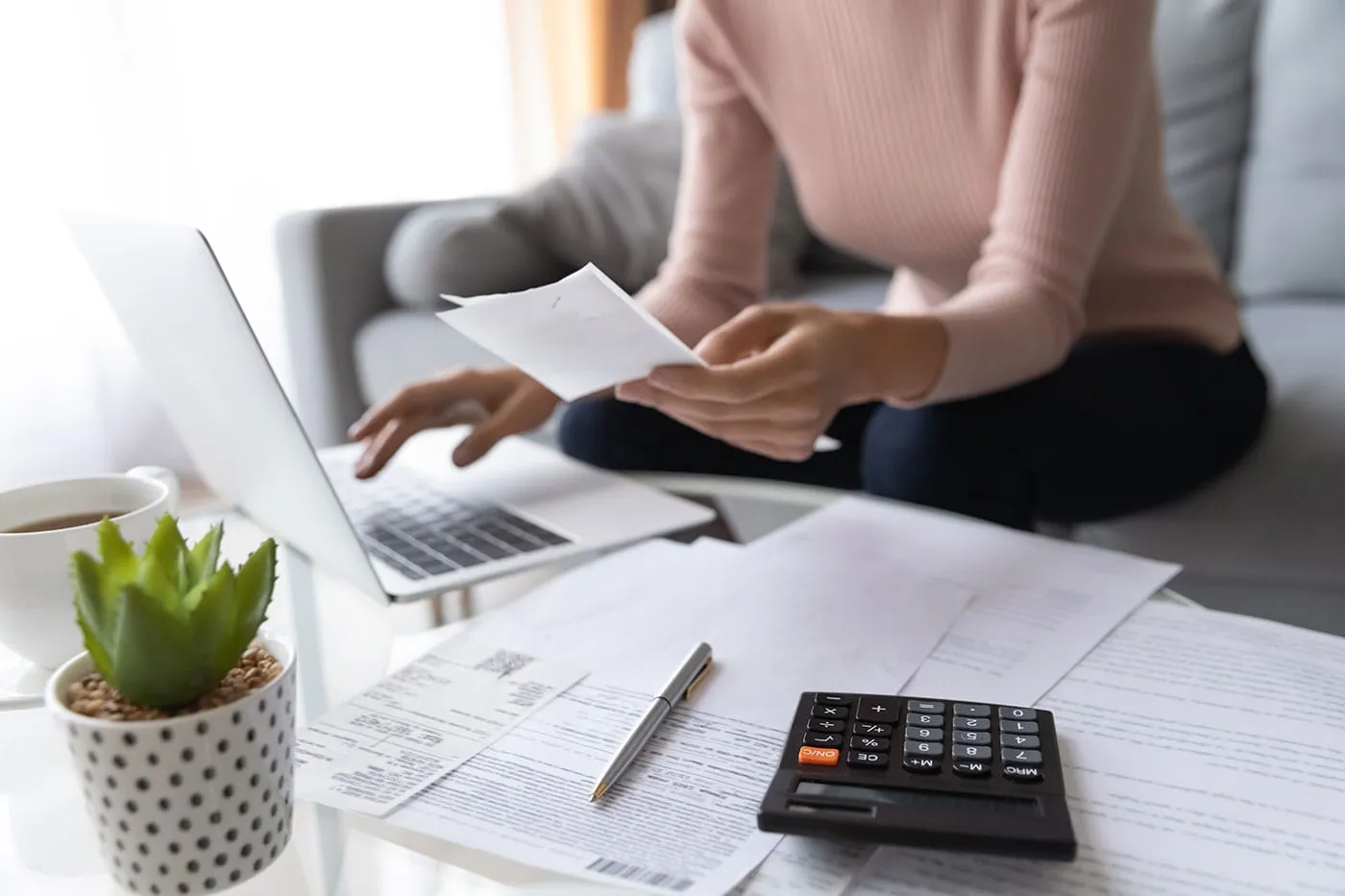 A woman sitting on the couch holds a receipt and uses her laptop while documents and a calculator sit on the table.