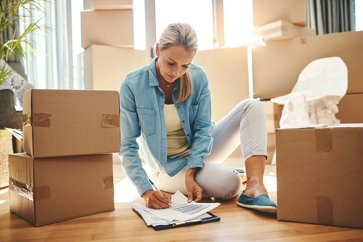 A woman signs documents on the floor of her home while brown moving boxes surrounds her.