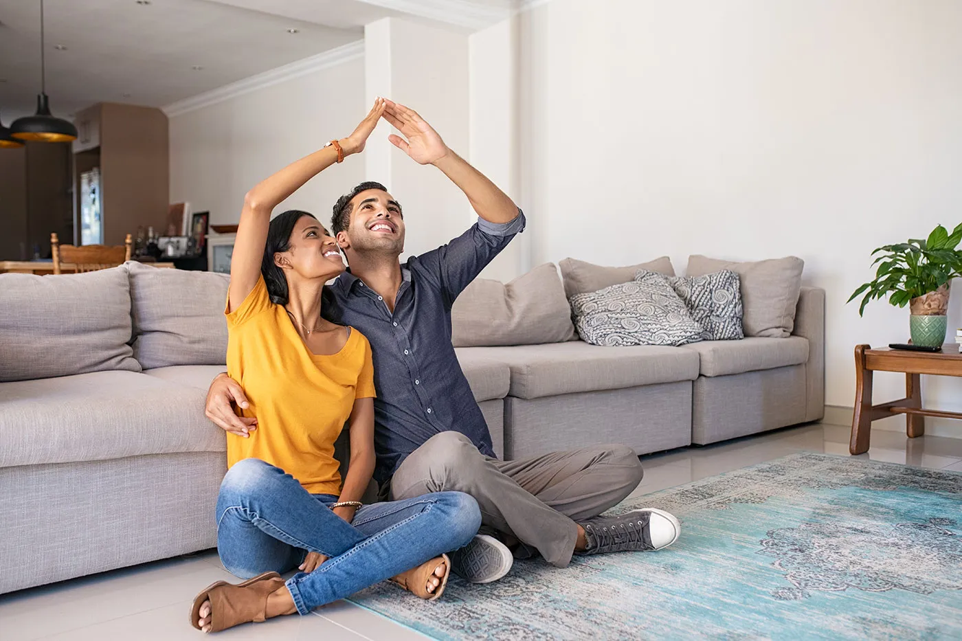 A couple sit on the ground of their living room as they connect their hands over their heads and smile.