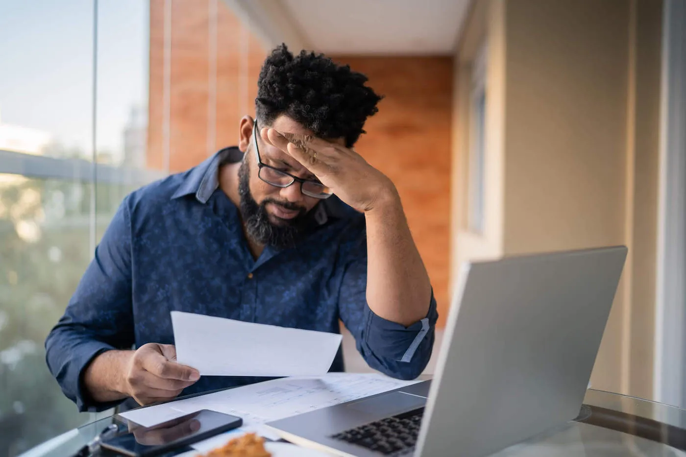 A man wearing a blue shirt has his hand to his forehead while looking at a document with his laptop on the table.