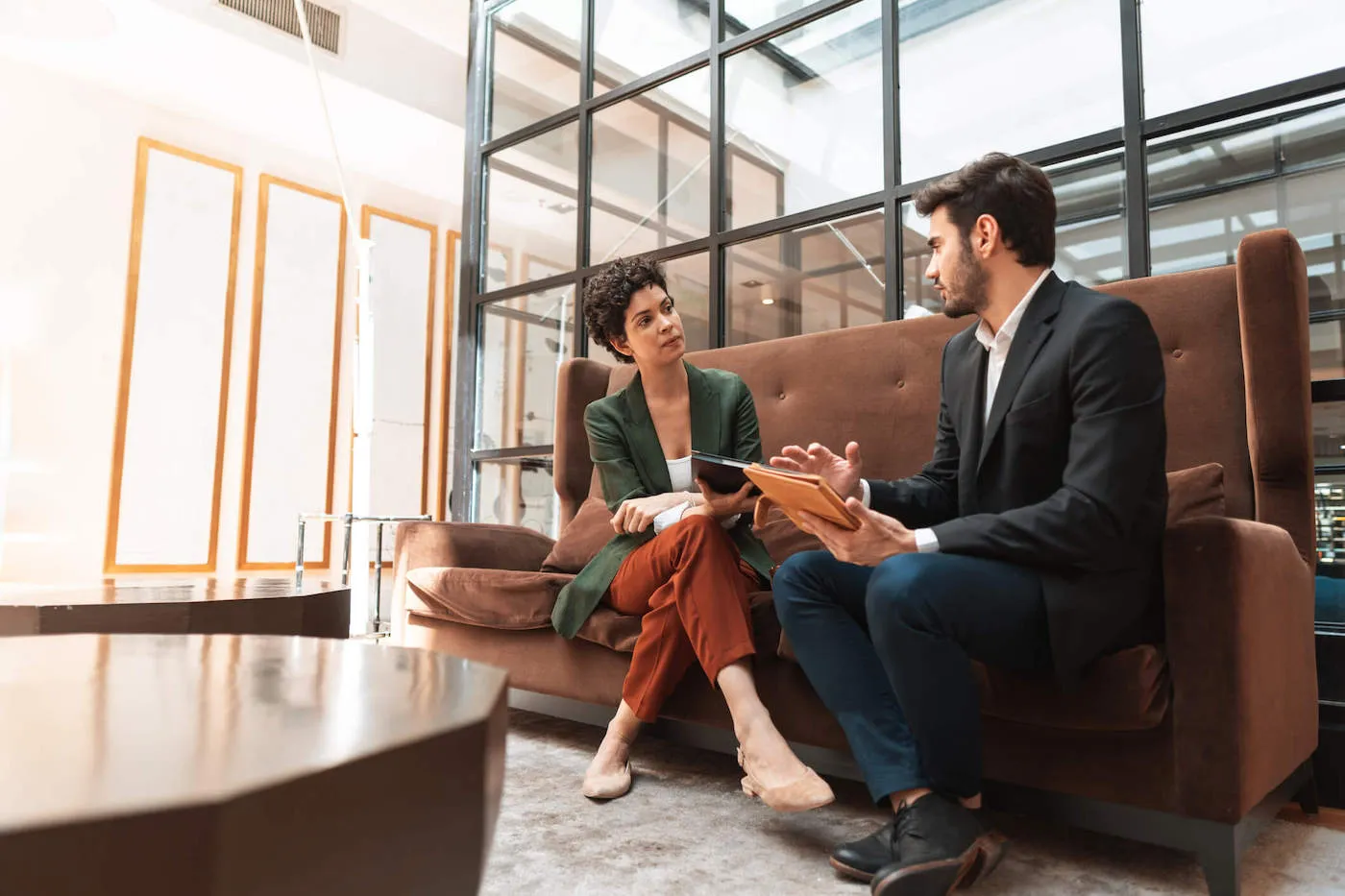 A man wearing a black suit talks with a woman across from him while they are both sitting on a brown couch.