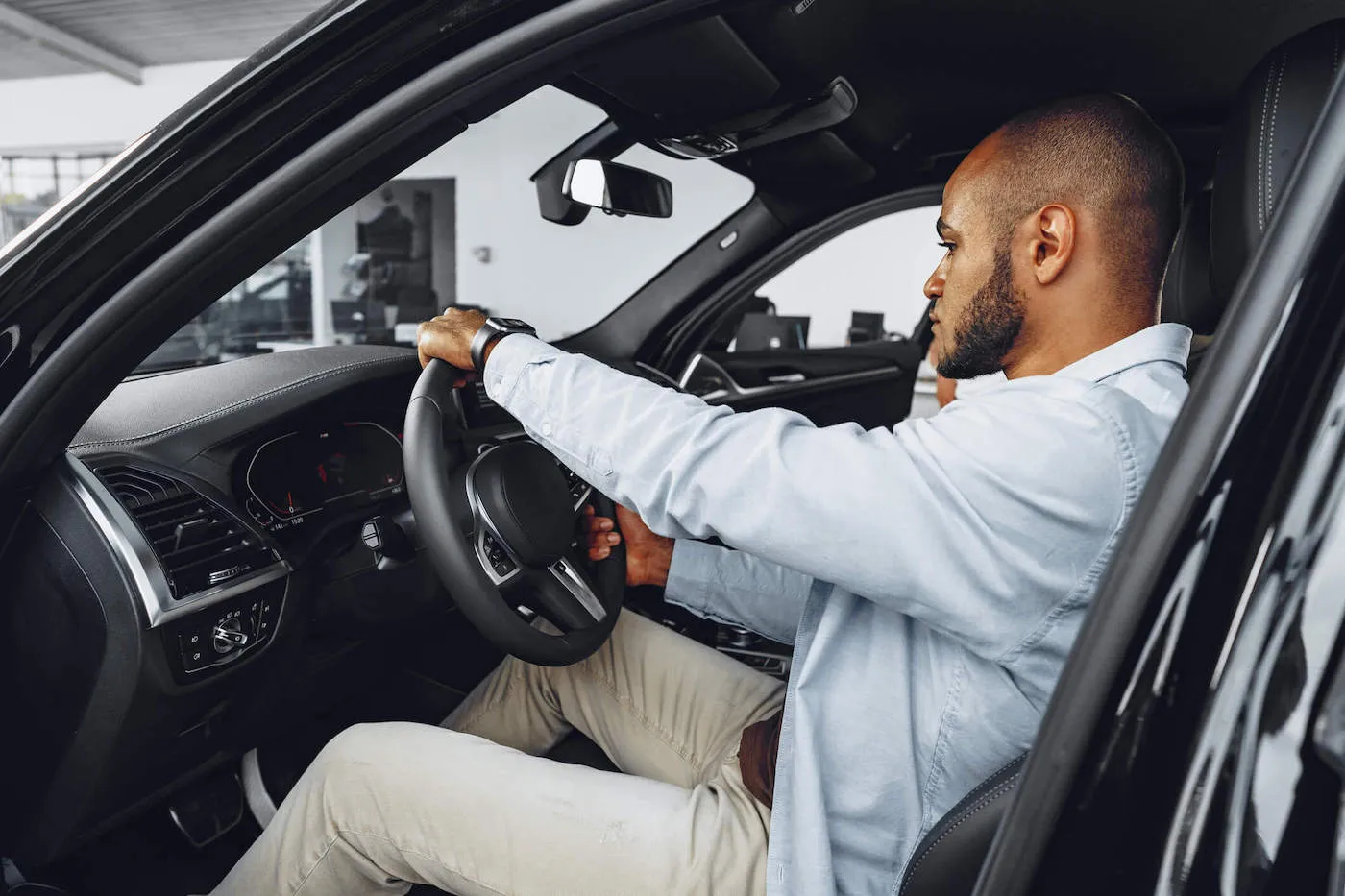 A man wearing a blue shirt inspects a car in the dealership while in the driver's seat.