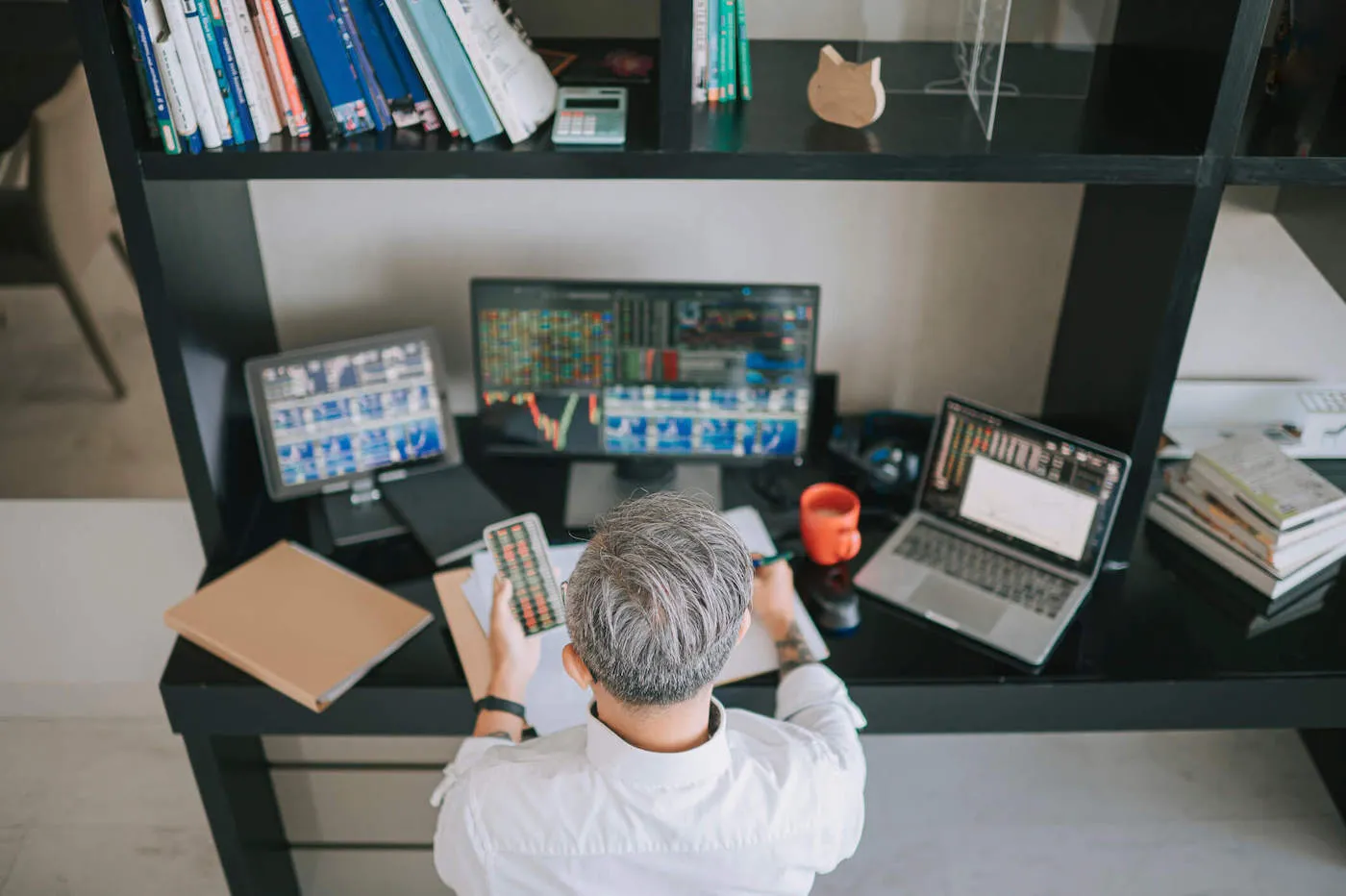 A man looks at stock prices on his phone while his other three computer screens on the table show other investment charts.