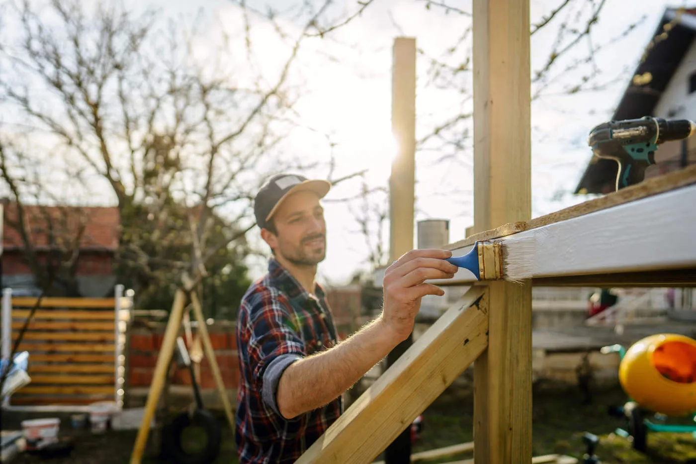 A man wearing a flannel shirt paints a piece of wood in his backyard white.