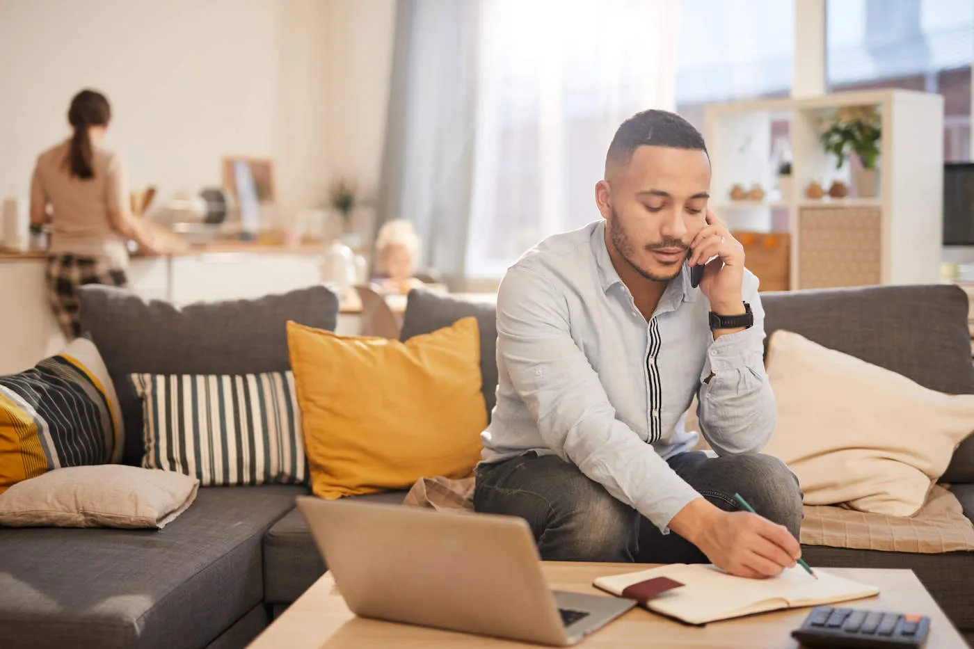 A man sits on his couch while talking on the phone and writing something down on his notebook.