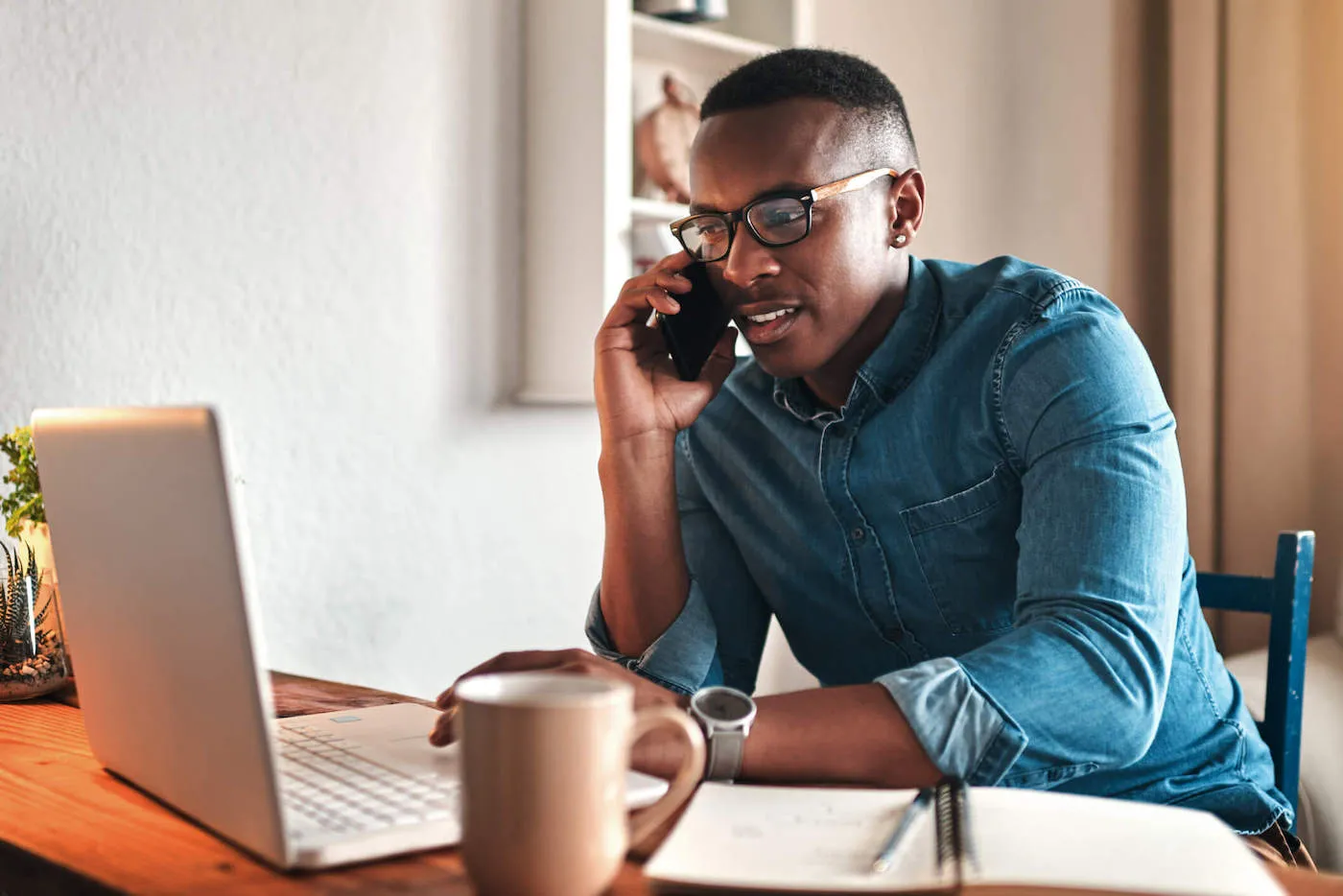 A man wearing a blue shirt is talking on the phone and working on his laptop at home.