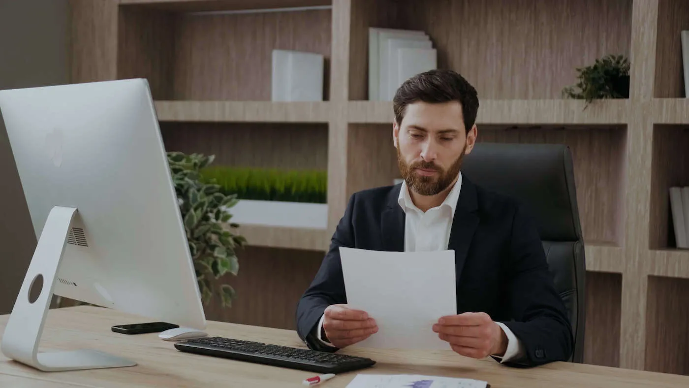 A man wearing a black suit jacket looks at a document from his work desk.