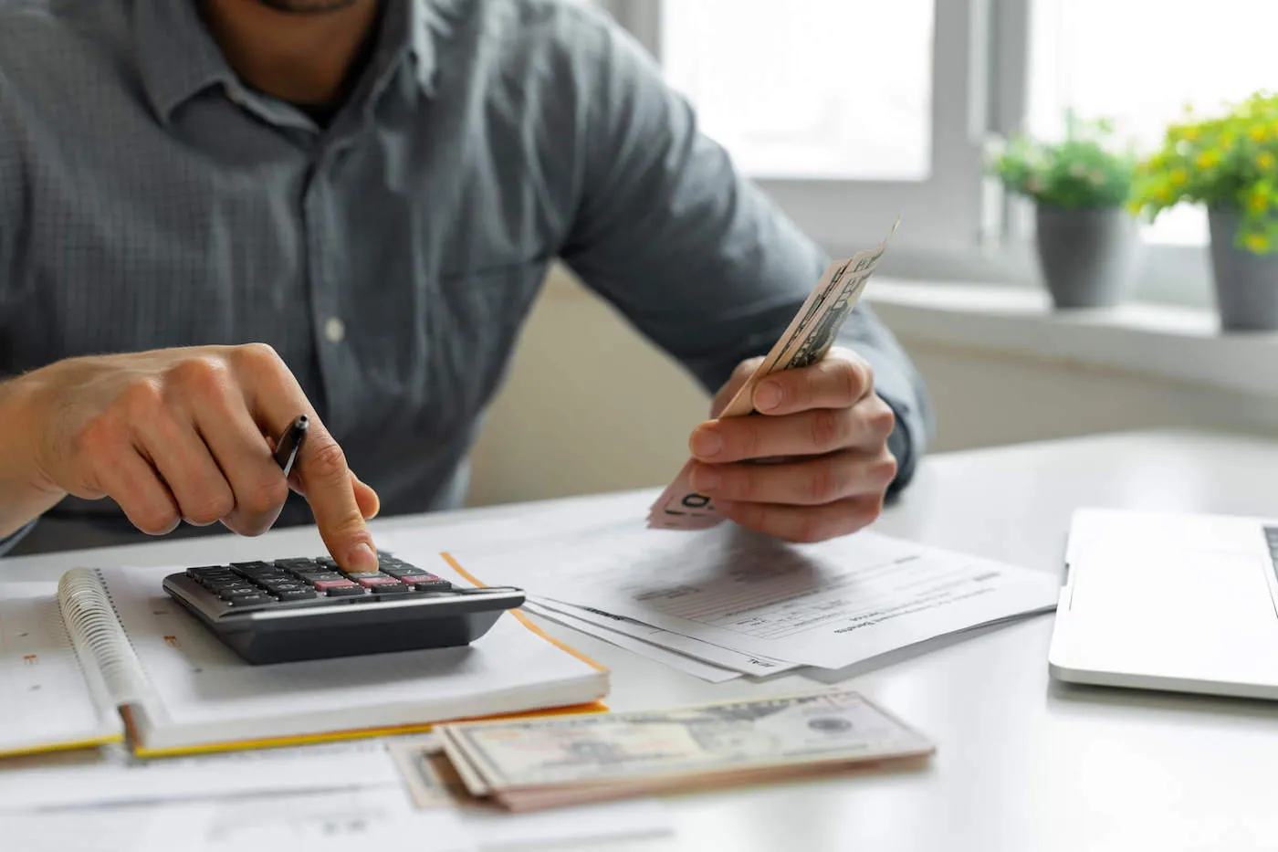 A man wearing a blue shirt is using a calculator while counting cash on the table.