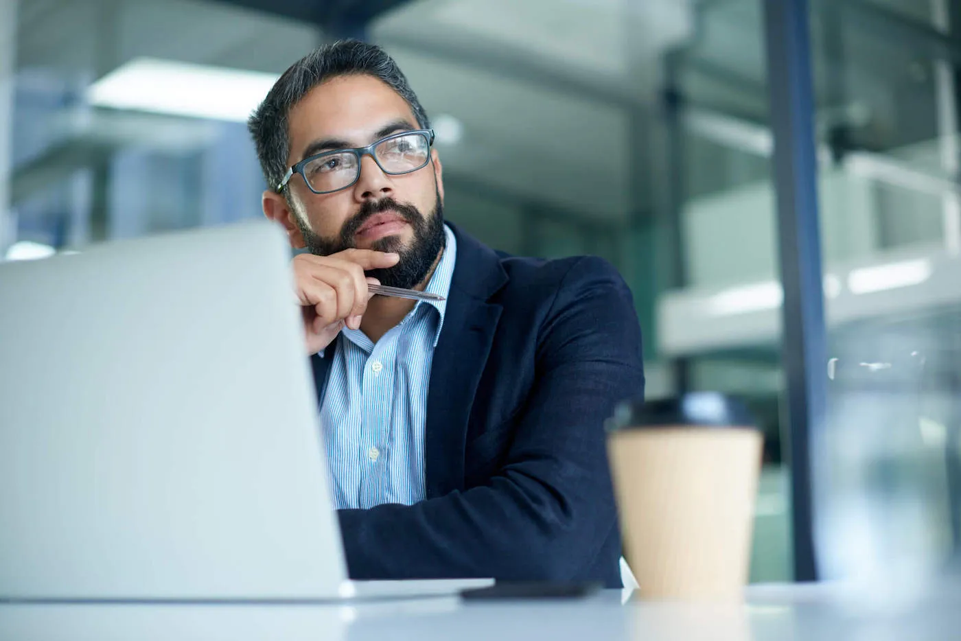 A man wearing a blue suit is looking out his office window while his laptop is in front of him.