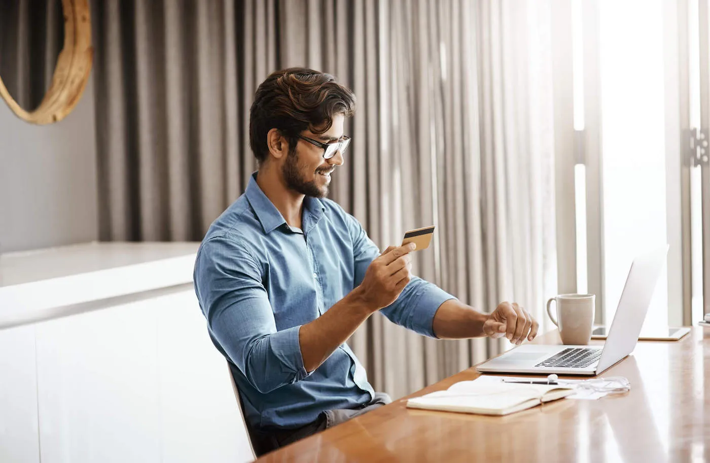 A man wearing a blue shirt and glasses smiles at his laptop while holding his credit card.