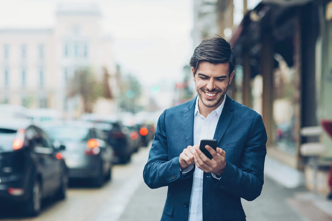 A man wearing a navy blue suit smiles at his phone while he is outside.