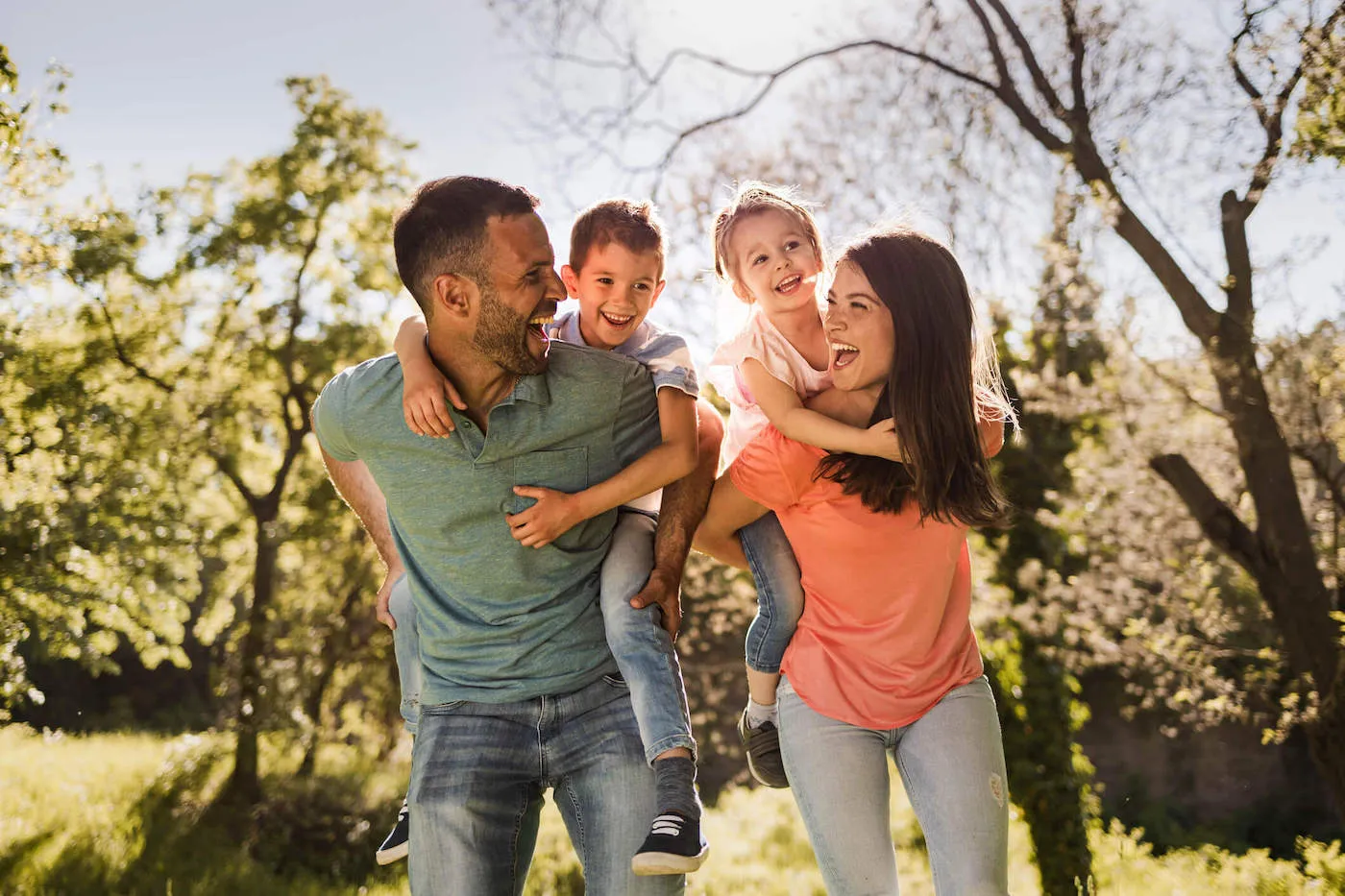 A mother and father smile at each other while giving their son and daughter piggyback rides outside.