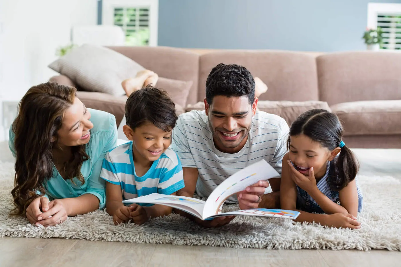 Mom and dad are reading a book to their two kids while lying down on the living room carpet.