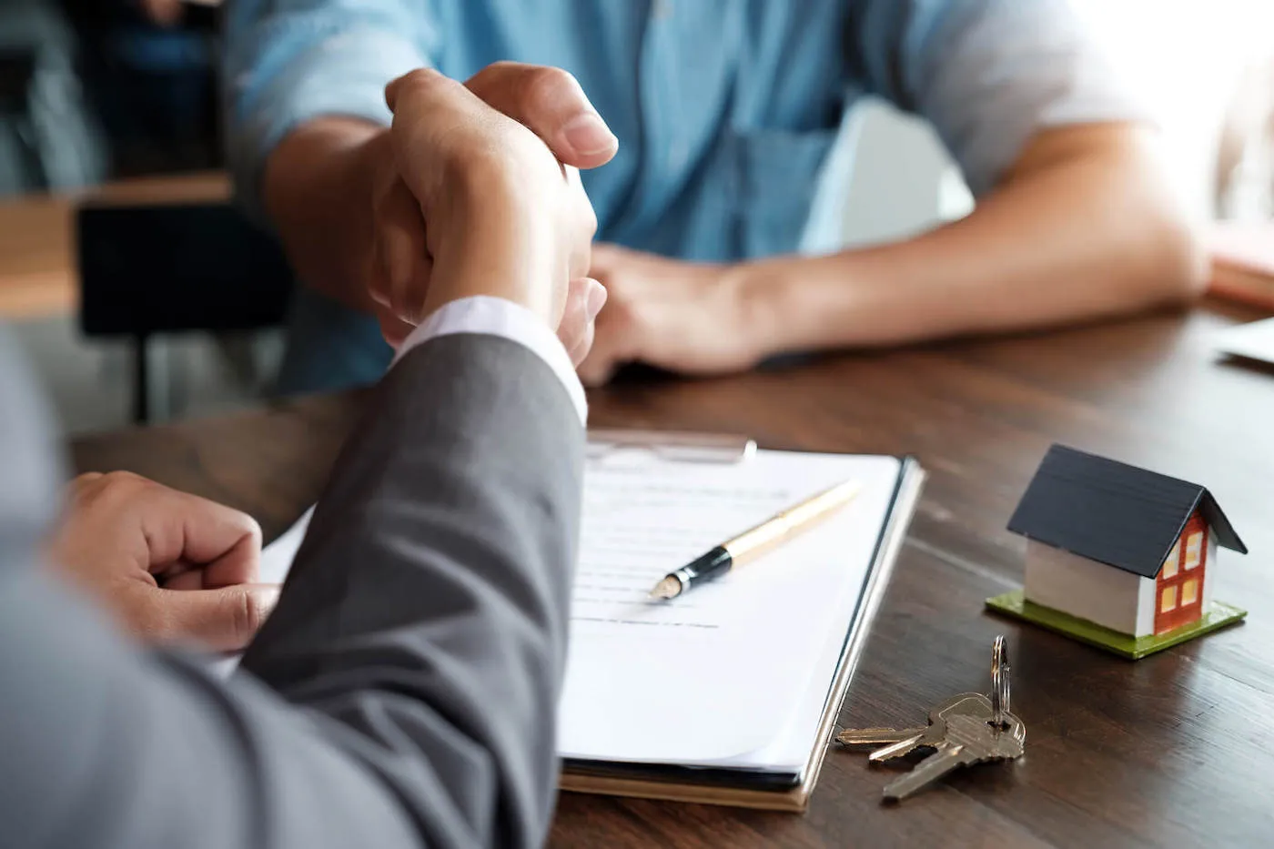 A person wearing a suit shakes hands with another person from across the table that has a document and house keys.