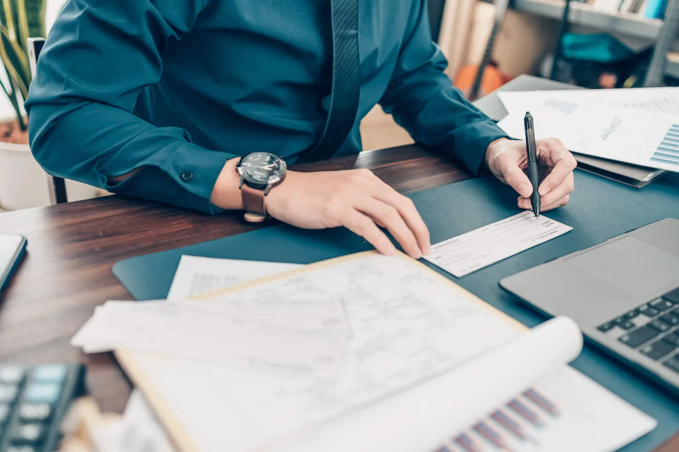 A person wearing a blue shirt writes on a check while having documents on their desk.