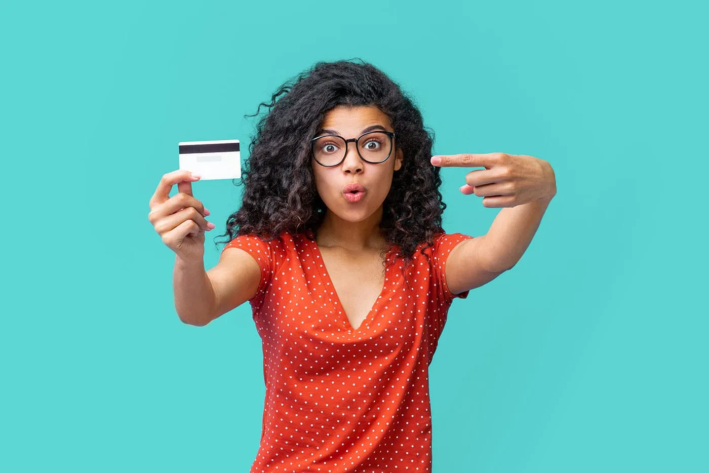 A woman has a surprised look on her face as she holds up her credit card and points at it with a light blue background behind her.