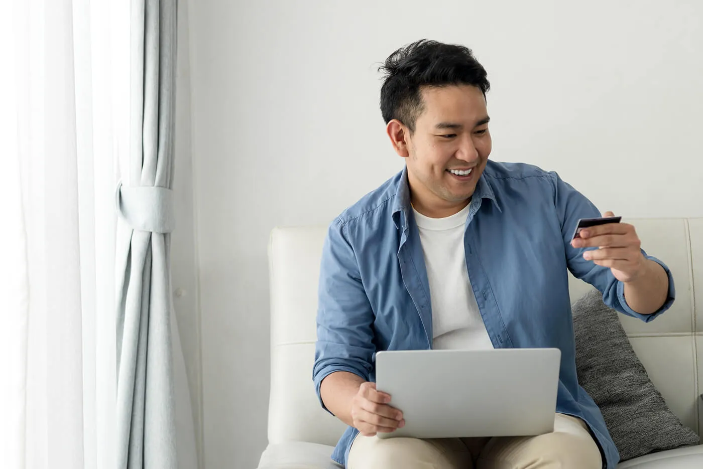 A smiling man holding a credit card and using a laptop while seated on a couch.