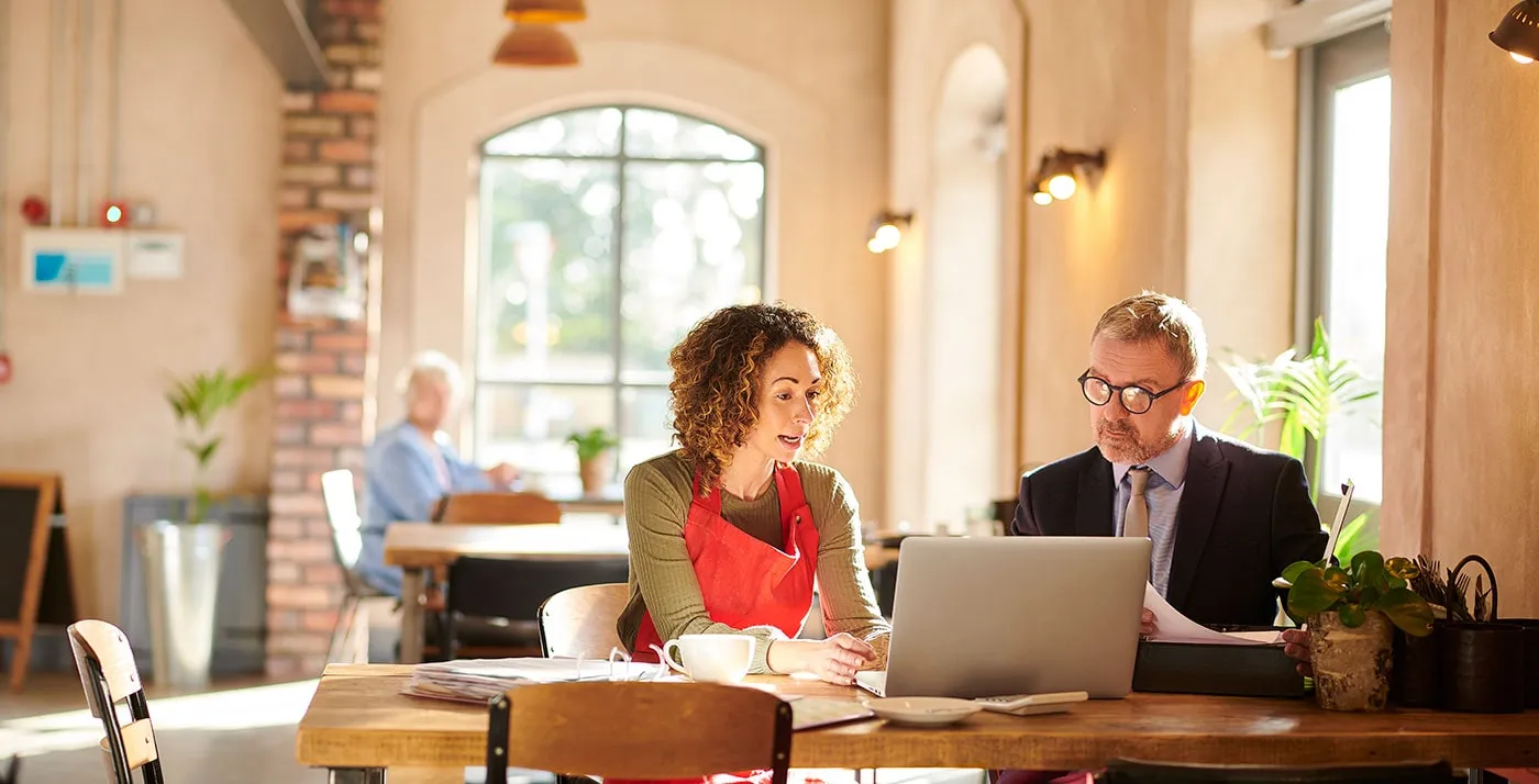 A business owner and a man wearing a suit talk to each other while looking at a laptop screen in front of them.