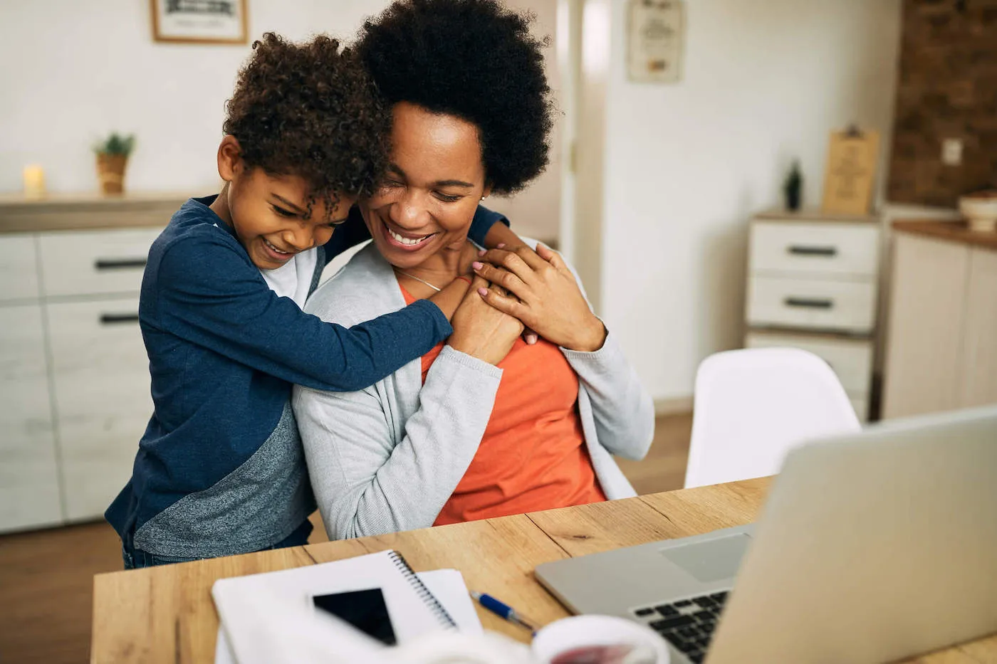 A mom and son hug each other while mom sits at the computer desk.