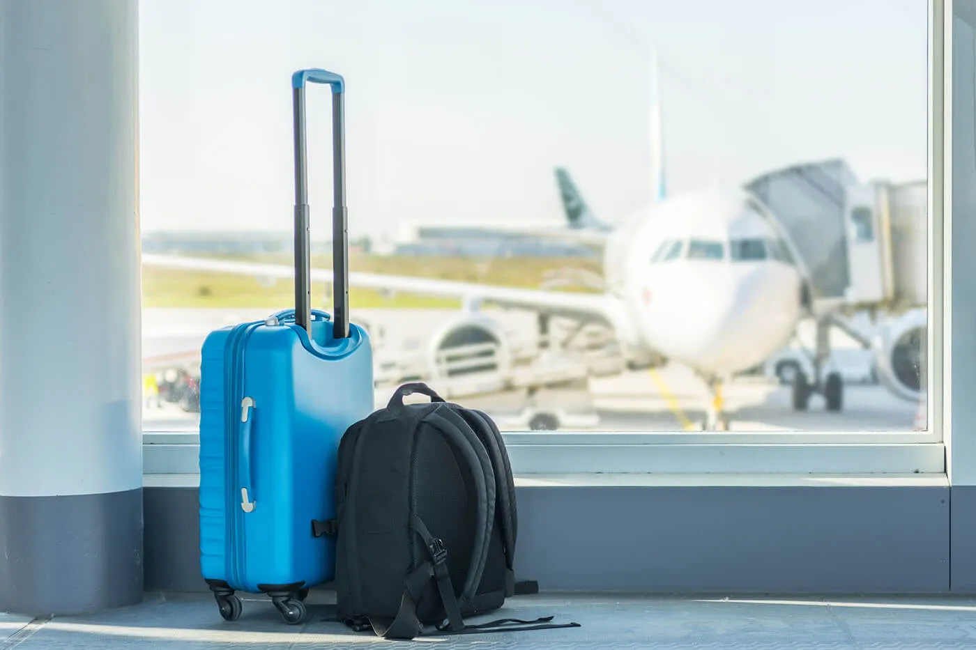 A bright blue roller suitcase and a black briefcase sitting on the floor of an airport terminal. An airplane is visible through the nearby window.