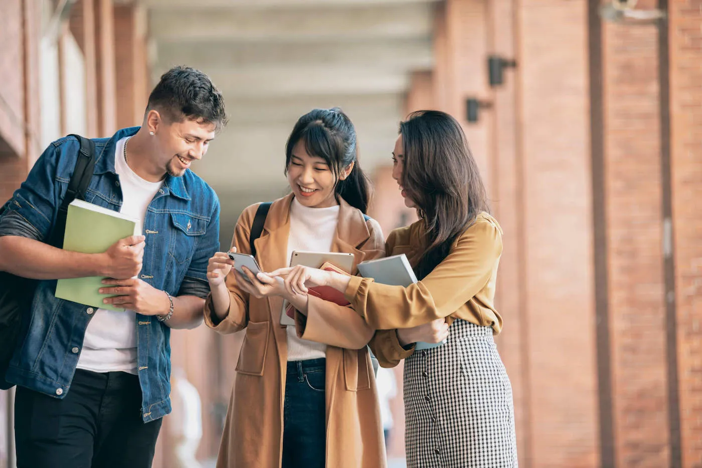 Three college students are laughing together while looking at a phone at their college campus.