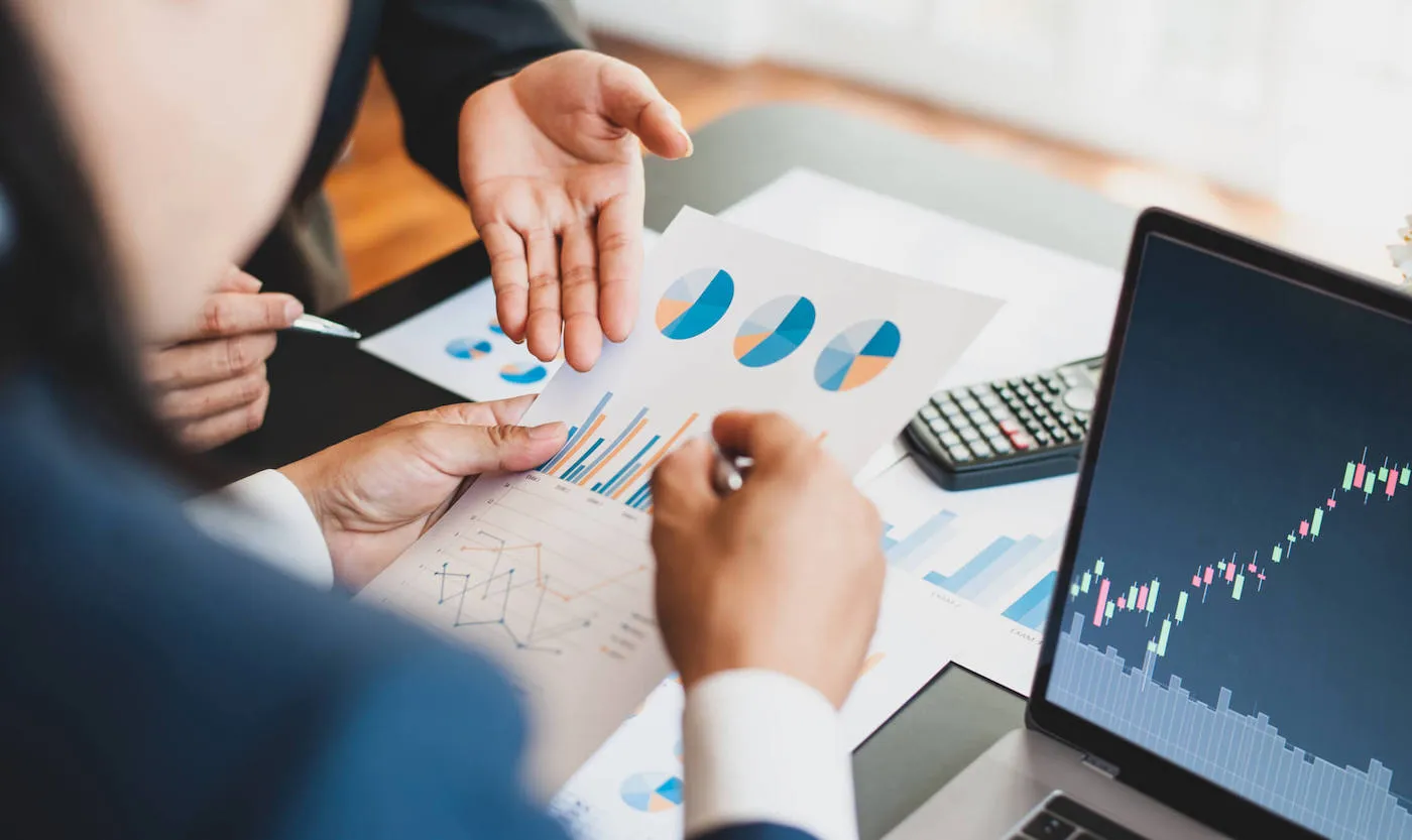 Two coworkers are looking at a document with graphs while a laptop shows a stock chart on the table.