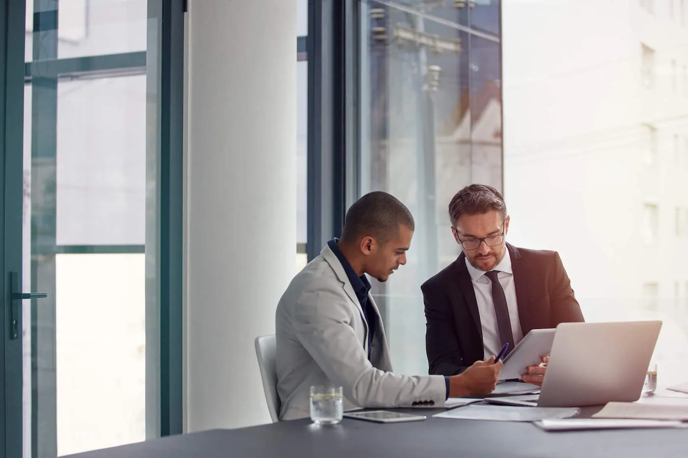 Two men wearing suits at the office are looking at a tablet while documents and a laptop are on the table.