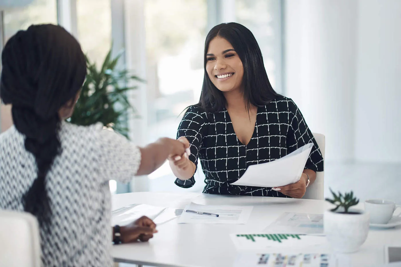 A woman wearing a black shirt smiles as she shakes hands with another woman across the table while holding a document.