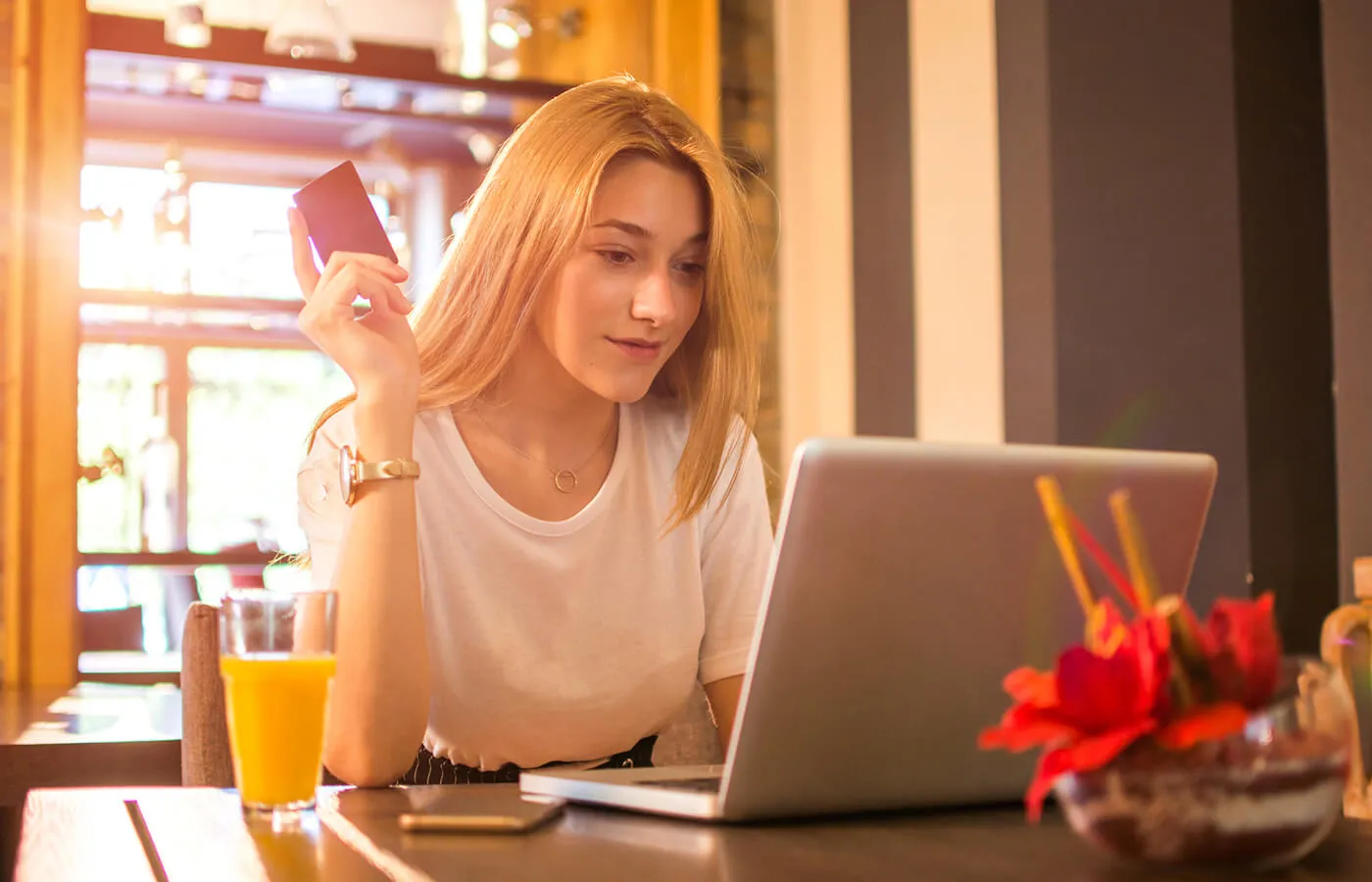 A woman with blonde hair sits down while holding her credit card and using her laptop on the table.