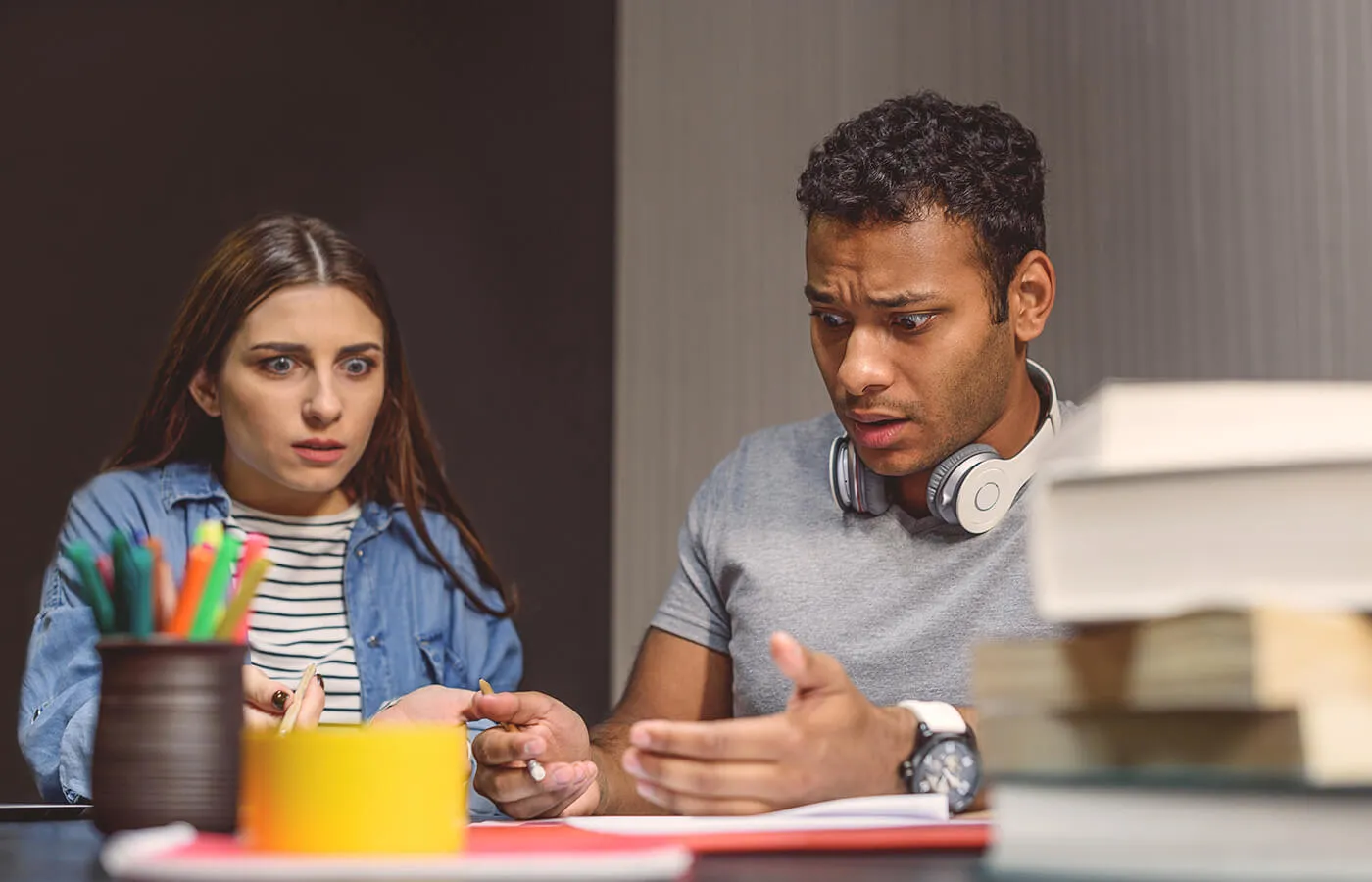 A young couple looks concerned and shocked while sitting at the desk with the books and a pencil holder