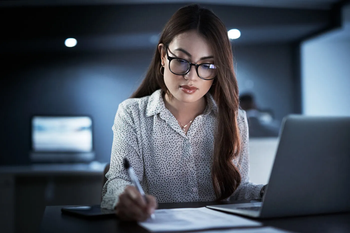 woman with black glasses writing on paper sitting on a desk with an open laptop