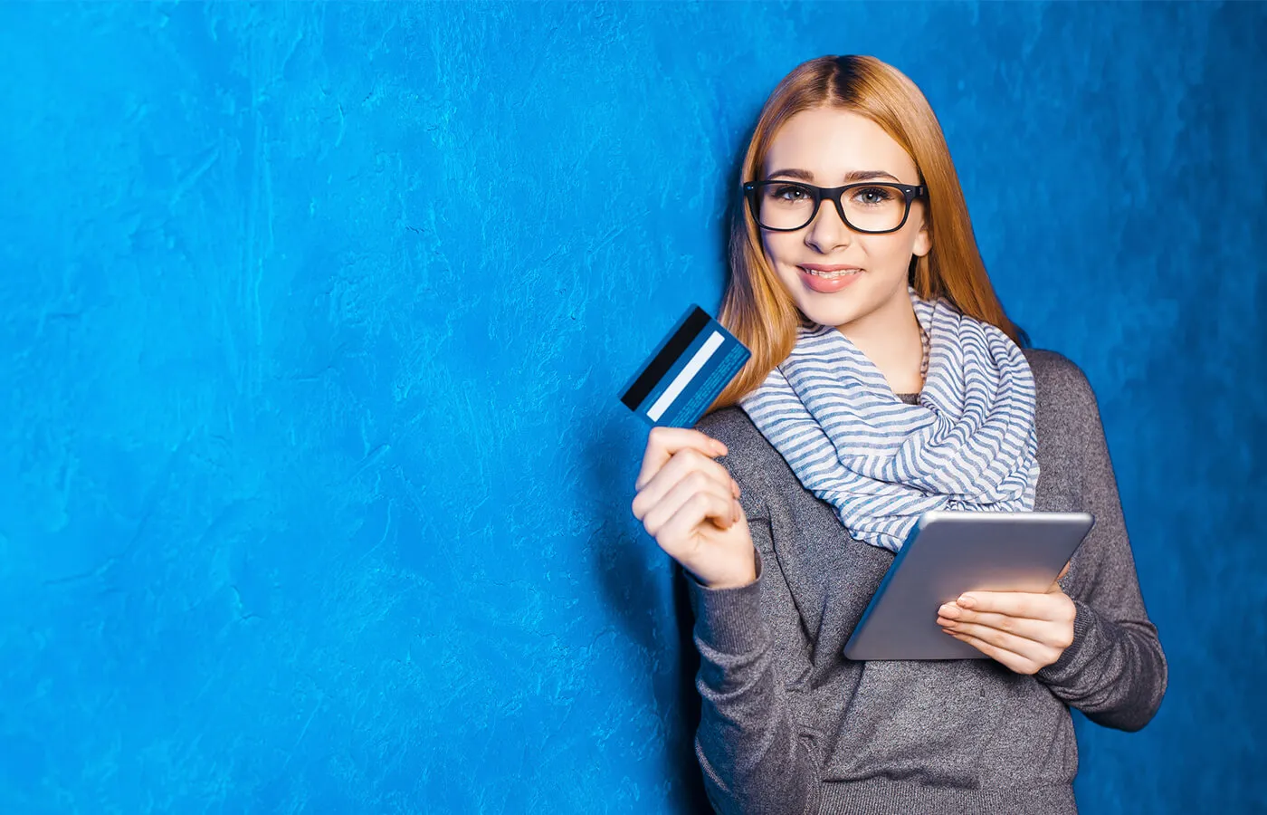 A woman standing against a blue background, holding her digital tablet in one hand and her credit card in the other.