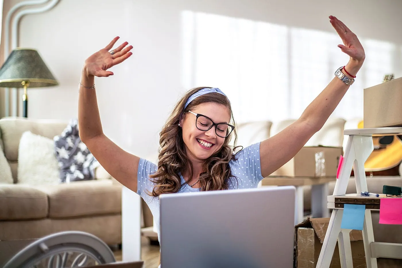 A woman wearing glasses and a polka dot shirt smiles as she raises her hands in the air with her laptop computer in front of her.