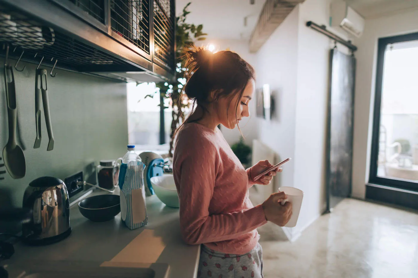 A woman holds a coffee mug while using her phone in the kitchen.