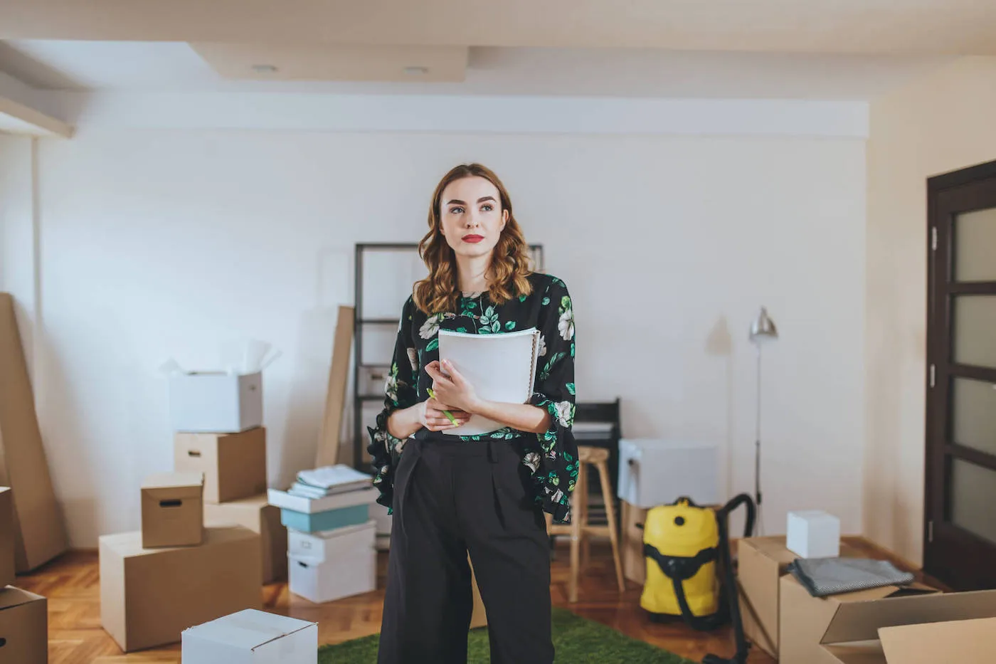 A woman holds her notebook while looking around her apartment that has moving boxes scattered around.