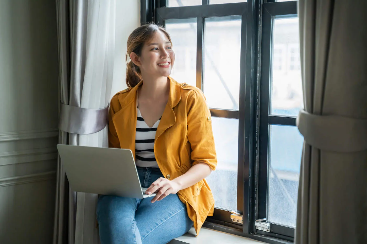 A woman wearing an orange jacket smiles while looking out the window and having her laptop on her lap.