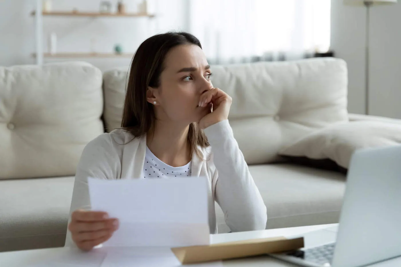 A woman frowns and has her hand to her mouth while looking out the window of her home and holding a document.