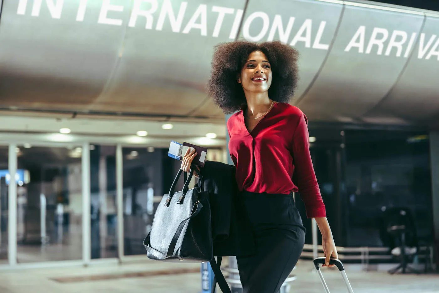 A woman wearing a red shirt smiles while carrying her luggage and ticket at the airport.