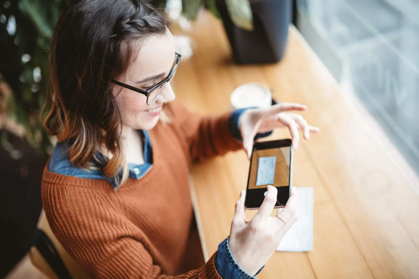 A woman wearing an orange sweater smiles while depositing her check by taking a picture of it through her phone.